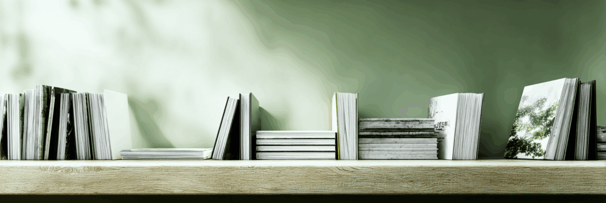 A wooden shelf lined with books and notebooks of varying sizes and textures. The background is softly shaded green, suggesting quiet growth and accumulated creative effort.