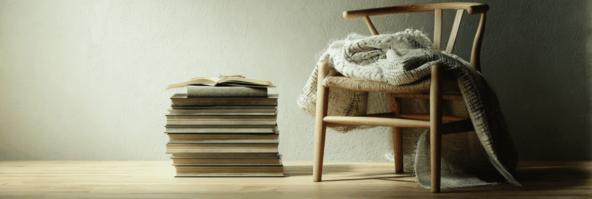 A woven chair draped with a light blanket sits beside a tall stack of books, topped with one open to a marked page. Gentle light and warm tones evoke rest, reflection, and the quiet weight of slow, meaningful work.