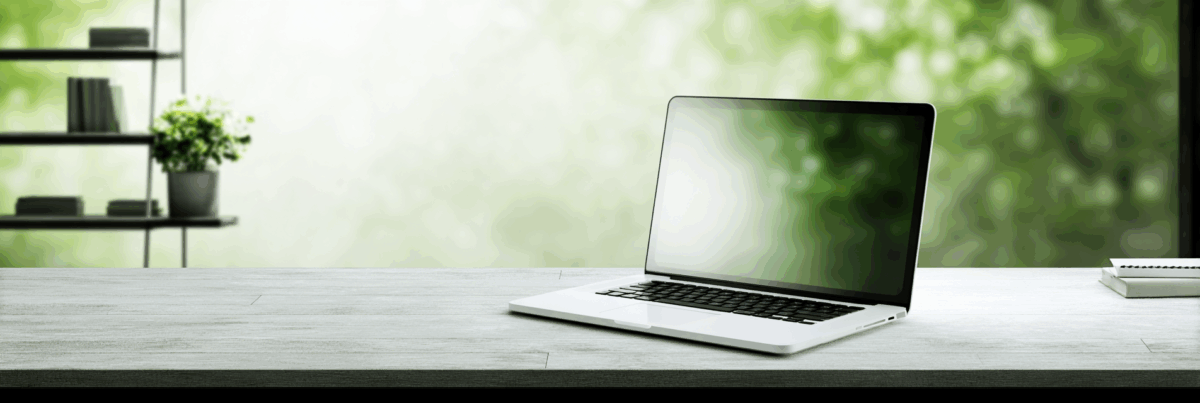 A softly lit workspace with a laptop on a concrete desk, its screen glowing faintly. A small plant rests on a stack of books, and a blurred lamp glows in the background. The mood is quiet and contemplative.