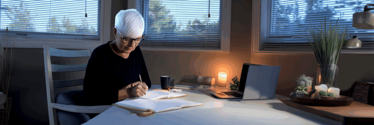 A white-haired woman sits at a softly lit desk, writing in a notebook beside a laptop and a glowing candle, surrounded by plants and quiet evening light filtering through window blinds.