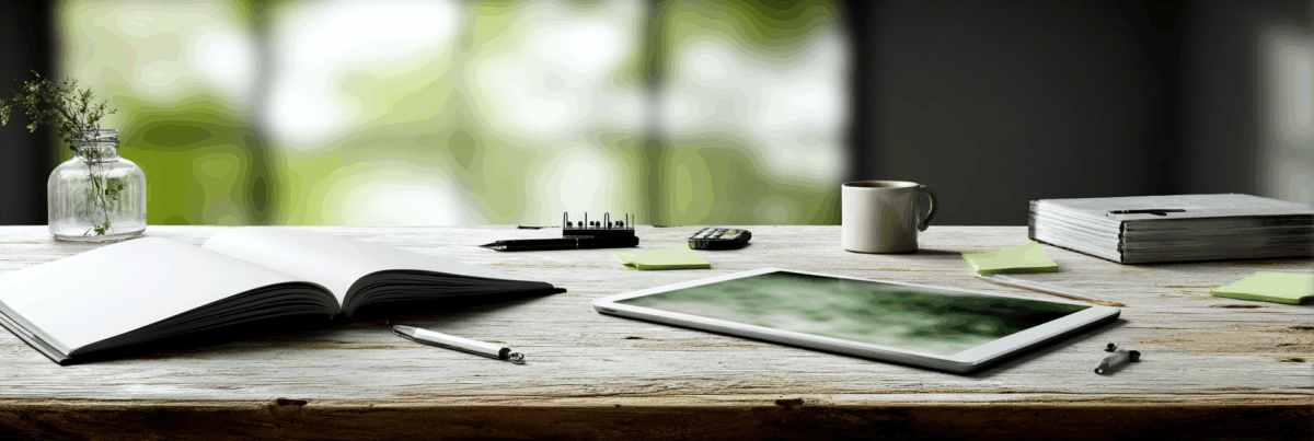 A rustic wooden desk with an open blank notebook, a silver pen, a digital tablet, and a beige coffee mug. A small glass vase with greenery sits to the left, with scattered sticky notes, a closed book, and a calculator in the background. Soft, blurred green light filters in from behind.
