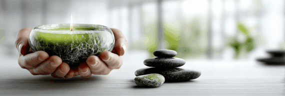 Close-up of two hands gently holding a green glass candle with a soft flame. Beside it, a small stack of smooth black stones rests on a light wooden surface, with blurred greenery in the background.