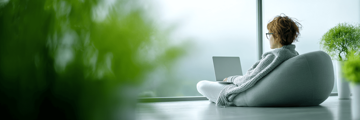 A person sitting in a soft chair near a large window, holding a laptop on their lap, with blurred greenery in the foreground and natural light filling the room.