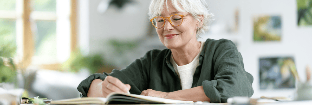 Older woman with short white hair and orange glasses smiles while writing in an open notebook at a bright, sunlit desk.
