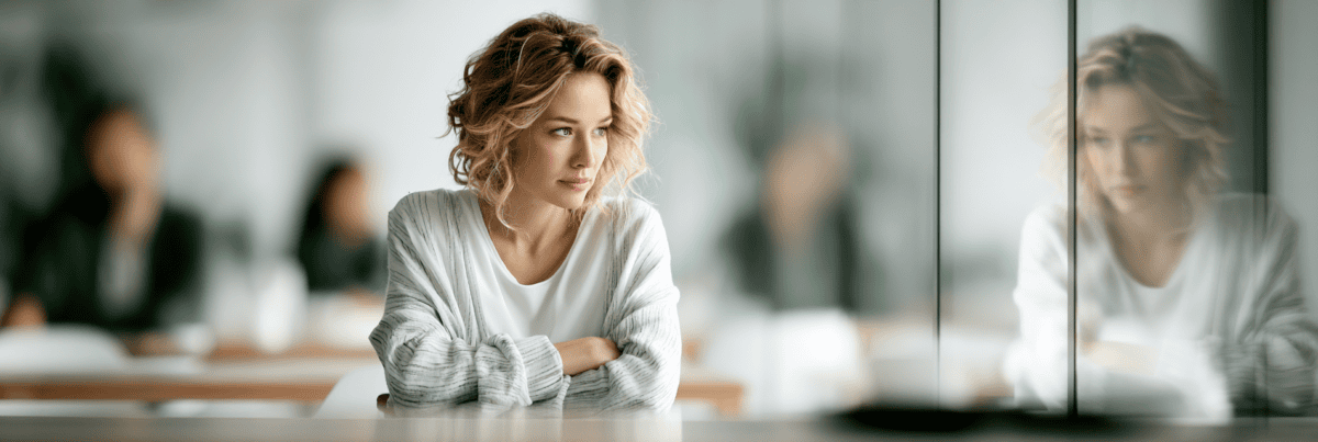 A thoughtful woman sitting in a calm, orderly room while her softened reflection appears in a glass wall beside her.